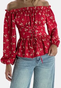 Blusa rossa floreale off-the-shoulder con maniche a sbuffo, vita cinchata e dettagli con lacci davanti, abbinata a jeans in denim blu a vita alta.