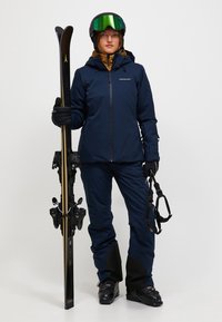Frau in navy Skijacke, Hose, Helm mit grünen Skibrillen, die Skier und Stöcke hält und schwarze Skischuhe trägt, vor einem einfarbigen Hintergrund.