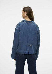 Jeans jakna v temno modri barvi, ki ima klasičen ovratnik, dolge rokave in gumbaste detajle na hrbtu. Teksturirana tkanina s sproščenim krojem.