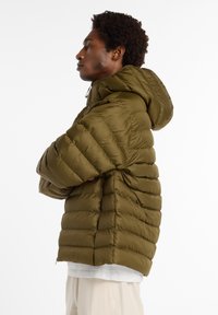 Veste rembourrée vert olive avec capuche amovible, présentant des sections matelassées et une fermeture éclair à l'avant. Portée par-dessus une chemise gris clair.
