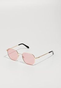Gafas de sol con montura de metal dorado, forma octagonal, lentes con tinte rosa y puntas de patilla negras. Almohadillas nasales transparentes realzan el diseño.