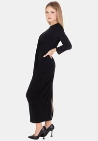 Abito nero a maniche lunghe con dettaglio a torsione in vita, spacchi laterali e una silhouette elegante, abbinato a scarpe nere con tacco alto.