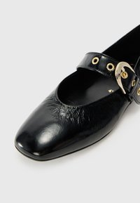 Scarpa flat nera in pelle Mary Jane con punta quadrata, cinturino laterale con hardware dorato e occhielli, e tomaia leggermente testurizzata.
