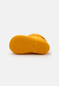 Semelle de botte en caoutchouc jaune avec un motif texturé et une forme arrondie. Présente une traction striée pour une bonne adhérence et une empreinte de logo de marque.