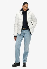 Hvid pufferjakke med quiltet design og sorte detaljer, parret med en marineblå turtle neck, lyseblå jeans og sorte snøresko.
