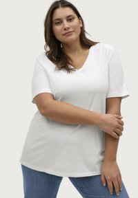 Hvid T-shirt med V-hals og korte ærmer, afslappet pasform, lavet af blødt stof; parret med blå jeans. Enkelt design og glat tekstur.