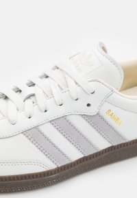 White leather sneakers with grey suede stripes, featuring a gold "SAMBA" label, white laces, and a textured brown rubber sole.