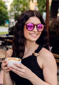 Sluneční brýle s růžovými reflektivními skly a želvovinovými rámy. Na sobě má strukturovaný černý top se zlatým akcentem. Drží bílý hrníček s kávou.