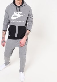 Šedá mikina s kapucí a velkým bílým logem Nike, černá přední kapsa a odpovídající šedé tepláky. Bílí tenisky doplňují celý outfit.