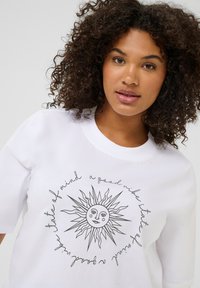 Sweatshirt blanc avec un graphisme de soleil dessiné à la main et du texte l'entourant. Encolure ronde et manches courtes. Texture de tissu douce.