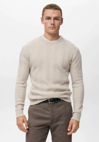Suéter de punto beige con un patrón de chevron texturizado, cuello redondo y mangas largas, combinado con pantalones marrón oscuro y un cinturón negro.