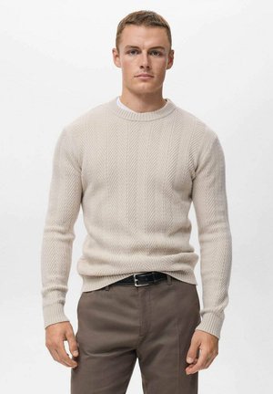 Suéter de punto beige con un patrón de chevron texturizado, cuello redondo y mangas largas, combinado con pantalones marrón oscuro y un cinturón negro.