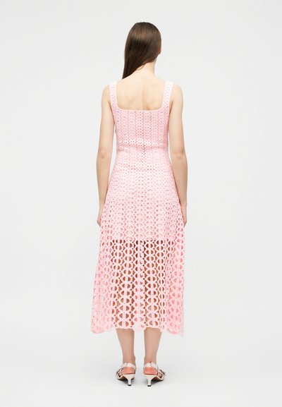 Femme aux longs cheveux bruns, vue de dos, portant une robe midi sans manches, rose à motifs, et des sandales blanches à talons.