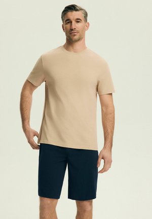 Muž oblečený v béžovom tričku s krátkym rukávom a tmavomodrých kraťasoch stojaci pred jednoduchým svetlým pozadím.