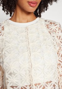 Vit blommig broderad topp med genomskinliga ärmar, texturerad tyg och ribbad krage med knäppning framtill.