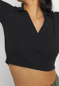 Musta lyhyt toppi, jossa on ribbineulos, V-pääntie ja lyhyet hihat, kauluskäällä ja viimeistelemättömät reunat.