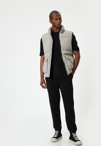 Lys grå polstret vest med høj krave, lynlåslukning og to frontlommer, parret med en sort t-shirt og sorte tapered bukser samt sorte sneakers.