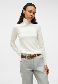 Cream turtleneck sweater with long sleeves worn with light gray jeans and a brown leather belt featuring a gold-tone buckle. Simple, fitted design.