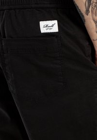 Pantalones negros con un bolsillo trasero visible, con una etiqueta de la marca blanca cosida en la cinturilla. El material parece suave y texturizado.