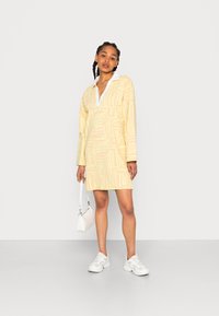 Vestido amarelo de mangas longas com colarinho branco, apresentando um design padrão abstrato. A modelo segura uma pequena mala branca e usa ténis brancos.