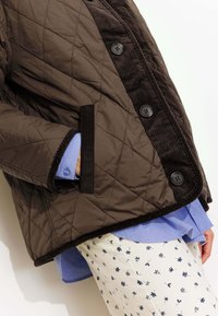 Veste matelassée marron avec des accents en velours côtelé noir, dotée de grands boutons et de poches latérales, portée sur une chemise bleu clair et un pantalon à motifs.