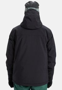 Schwarze isolierte Jacke mit Kapuze, die eine glatte Textur und elastische Bündchen aufweist, kombiniert mit türkisfarbenen Hosen, von hinten betrachtet.