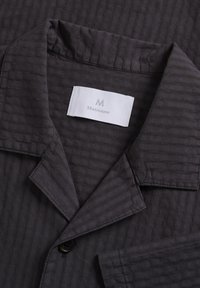 Veste en tissu gris foncé texturé avec col à revers cranté, bouton noir, et étiquette blanche portant l'inscription "Matinique" à l'intérieur du col.