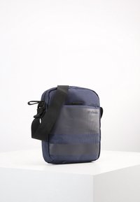 Borsa a tracolla blu navy realizzata in tessuto texturizzato, con pannello frontale grigio, tracolla regolabile e dettaglio del logo. Design leggero e compatto.