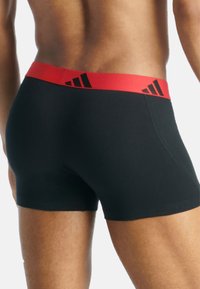 Μαύρα μποξερ σλιπ με κόκκινη ελαστική μέση που διαθέτει μια μαύρη μοτίβο λογοτύπου. Το ύφασμα φαίνεται λείο και κομμένο.