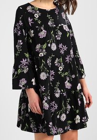 Czarna sukienka w kwiaty z rękawami o długości trzy czwarte w kształcie dzwonka, zdobiona purpurowymi i białymi wzorami kwiatowymi, z falbaniastym dołem i luźnym krojem.