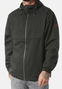 Giacca verde scuro con zip, cappuccio, polsini elasticizzati e tasca sul petto. Presenta una toppa con logo sulla manica sinistra e una texture liscia.