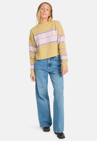 Gehaakte gele trui met paarse en roze strepen, ronde halslijn en een relaxed fit. Gecombineerd met hoge blauwe wijdere jeans.