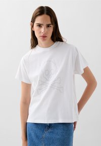 Bílý bavlněný tričko s kulatým výstřihem, na kterém je stříbrný design lebky a zkřížených kostí v grafickém stylu. Uvolněný střih s krátkými rukávy.