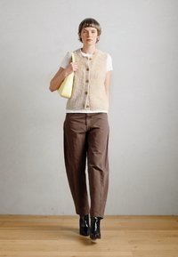 Bežna pletena jopica z gumbom spredaj, bela majica, rjave hlače in črne gležnjarje. Drži majhno rumeno torbico.