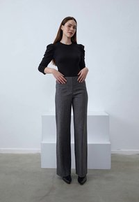Femme portant un haut noir à manches longues et un pantalon large taille haute gris, se tenant devant des blocs blancs en escalier sur un sol gris.