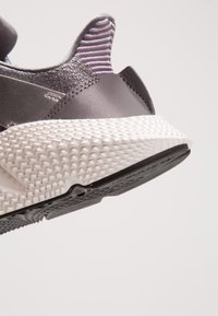 Grå sneakers med en texturerad vit mellansula och svart yttersula, med en slät läderöverdel och en lila randig accent vid kragen.