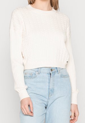 Woman wearing a white cable-knit sweater with ribbed sleeves and light blue high-waisted jeans, standing against a plain background.