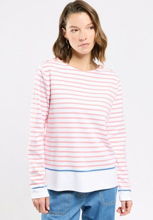 Femme aux cheveux bruns mi-longs portant une chemise blanche à manches longues avec des rayures roses et un ourlet bordé de bleu, et un jean bleu.