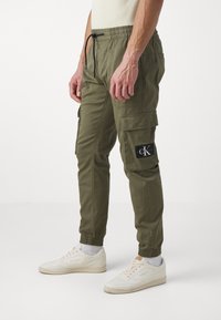 Olivově zelené cargo kalhoty s elastickým pasem, nastavitelnou šňůrkou, bočními kapsami a černým logem na levém stehně.
