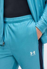 Άτομο που φοράει ανοιχτό μπλε παντελόνι αθλητικής φόρμας Under Armour και ταιριαστό μπουφάν, με το χέρι στην τσέπη, και μαύρο πουκάμισο ορατό από μέσα.