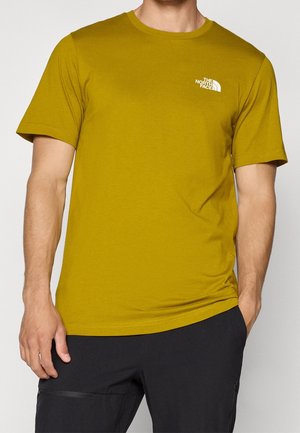 Mann trägt ein kurzärmeliges Senfgelbes T-Shirt mit The North Face Logo auf der Brust und schwarze Hosen mit Taschen.