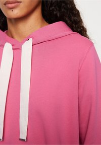 Roze hoodie met een trekkoord van witte stof, zachte textuur en een relaxte pasvorm, met nadruk op de capuchon en stikdetails.