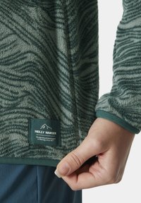 Veste en polaire verte avec un motif ondulé, dotée d'une étiquette, de poignets élastiques et d'une ouverture zippée à l'avant. Texture douce, conçue pour la chaleur.