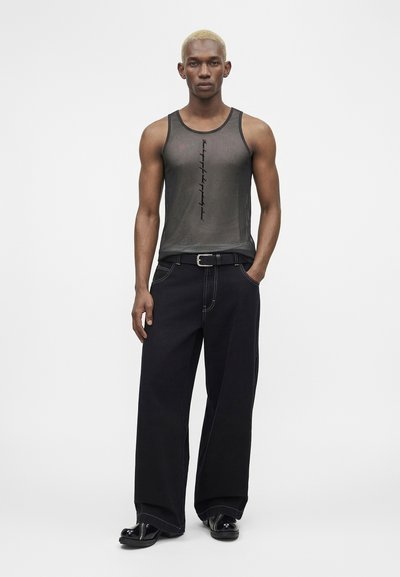 Άνδρας με κοντά ξανθά μαλλιά φοράει μαύρο διάφανο αμάνικο με κάθετο κείμενο, φαρδιά μαύρα παντελόνια, μαύρη ζώνη και μαύρα παπούτσια, στέκεται ακίνητος.