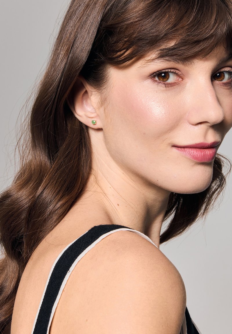 Złote kolczyki wkrętki z zielonym kamieniem szlachetnym, ramię modelki widoczne, ciemne włosy w delikatnych falach, ubrana w czarną bluzkę z białym paskiem.