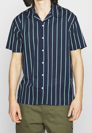 Mann trägt ein marineblaues Kurzarm-Hemd mit weißen und grünen vertikalen Streifen und olivgrüne Hosen.