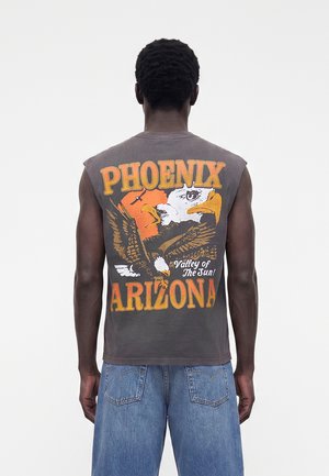 Person trägt ärmelloses dunkelgraues Shirt mit Adlergrafik und orangefarbenem Text "Phoenix Arizona Valley of the Sun", kombiniert mit blauen Jeansshorts.