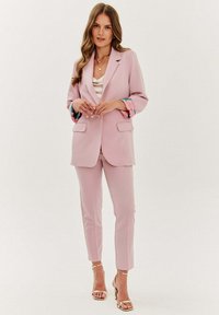 Růžový blejzer se dvěma předními kapsami a hlubokým výstřihem, doplněný o ladící strukturované kalhoty. Manžety mají květinový vzor.