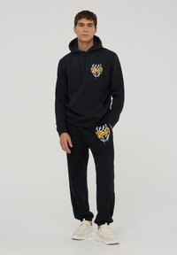 Czarna bluza z kapturem i spodnie dresowe wykonane z miękkiego materiału. Bluza ozdobiona grafiką w bieli i żółci. Spodnie mają dopasowaną grafikę.