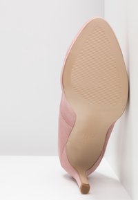 Escarpin en daim rose à talon haut avec bout pointu, semelle inclinée montrant une semelle texturée et talon fin appuyé contre des surfaces blanches.